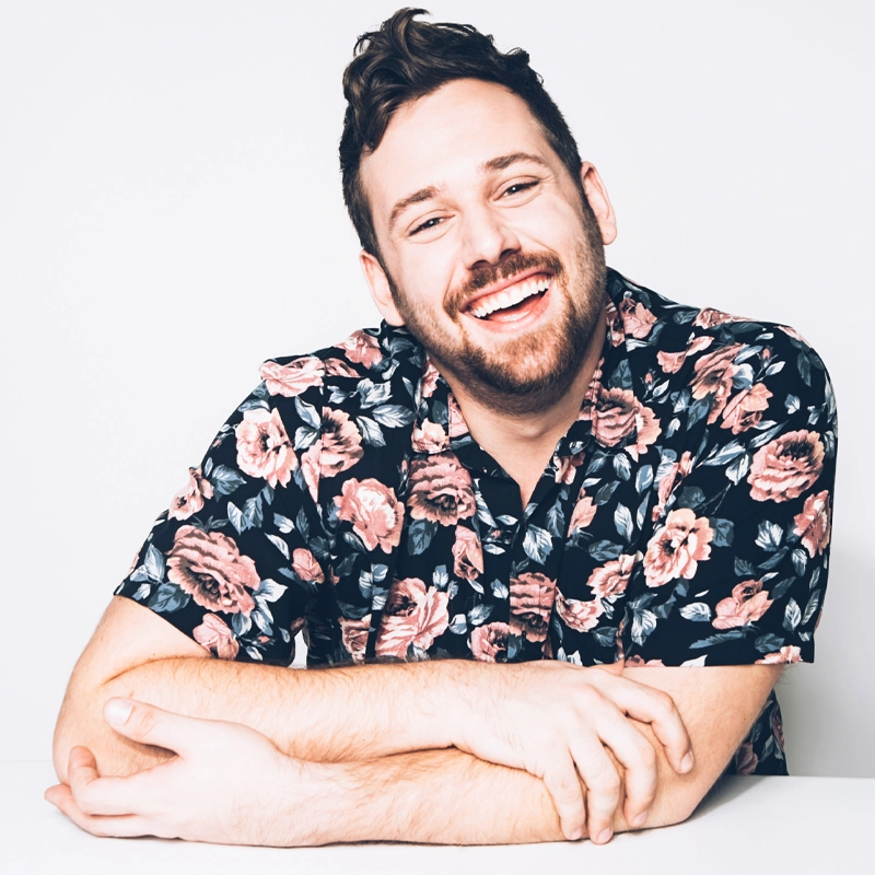 A smiling man with short brown hair and a beard sits at a white table, arms folded. He wears a black shirt with a pink floral pattern. The background is plain white, giving the image a bright and cheerful feel.