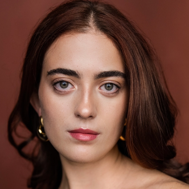 A woman with fair skin and long, wavy brown hair gazes directly at the camera. She has thick eyebrows, green eyes, and wears gold hoop earrings. The background is a warm reddish-brown color. Her expression is calm and neutral, embodying the poised elegance of Mairéad O’Neill.