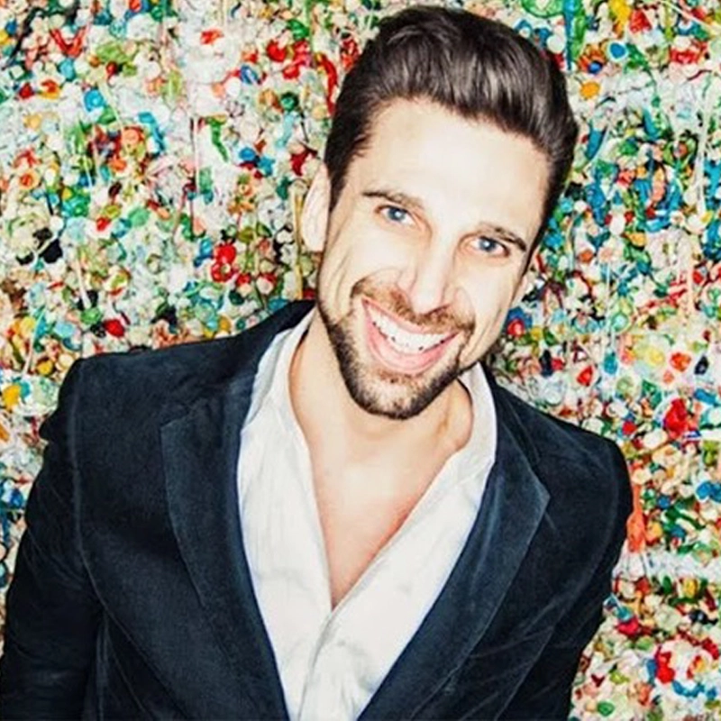 A smiling man with short brown hair and a trimmed beard stands in front of a colorful wall covered with chewed gum. He wears a black jacket over a white shirt and looks directly at the camera, tilting his head slightly.