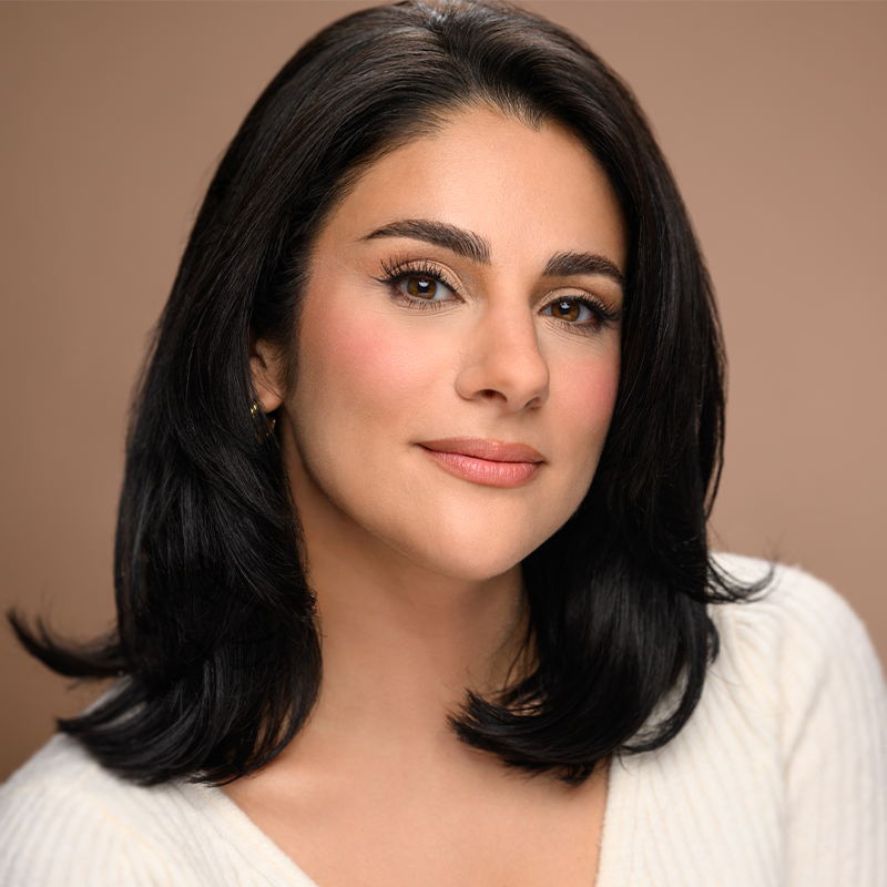 Gianna Yanelli, with medium-length glossy black hair and light skin, looks confidently at the camera. She wears natural makeup with subtle blush and nude lipstick, a white ribbed top, and small gold hoop earrings against a soft tan background.