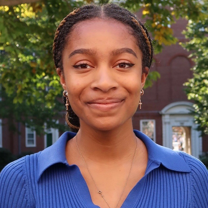 Gabrielle Greene, a young woman with braided hair, smiles gently outdoors in bright sunlight. She wears a blue collared top, small hoop earrings with cross charms, and a necklace. Green trees and a red brick building stand behind her.