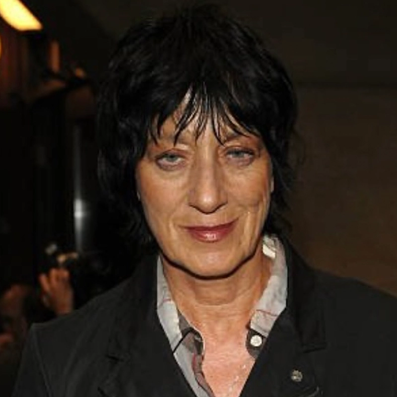Katherine Winter, with short, tousled black hair, light skin, and blue eyes, wears a dark blazer over a light shirt. She smiles slightly against a dim background where another figure is blurred and a camera flash is visible.