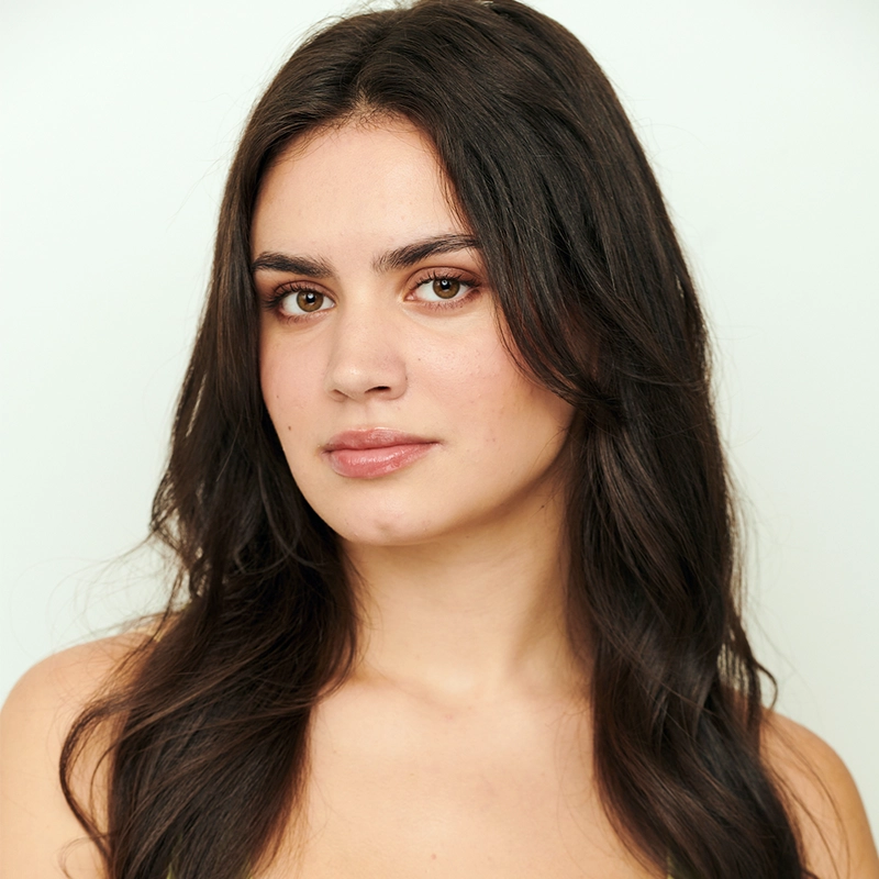 Katherine Winter, a young woman with long, wavy dark brown hair and light skin, looks calmly at the camera. She has natural makeup and thick eyebrows, standing against a plain, light-colored background with a neutral, relaxed expression.