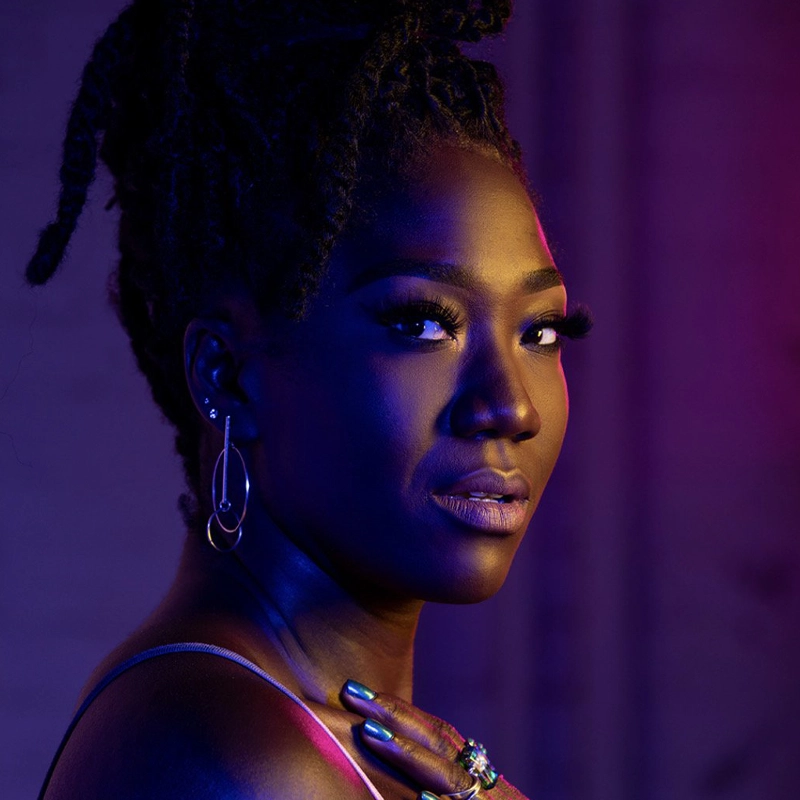 A woman with dark skin and long, textured hair styled up gazes over her shoulder. She wears hoop earrings, rings, and a strappy top. Blue and purple lighting creates dramatic shadows on her face and body, giving the portrait an artistic, moody feel.