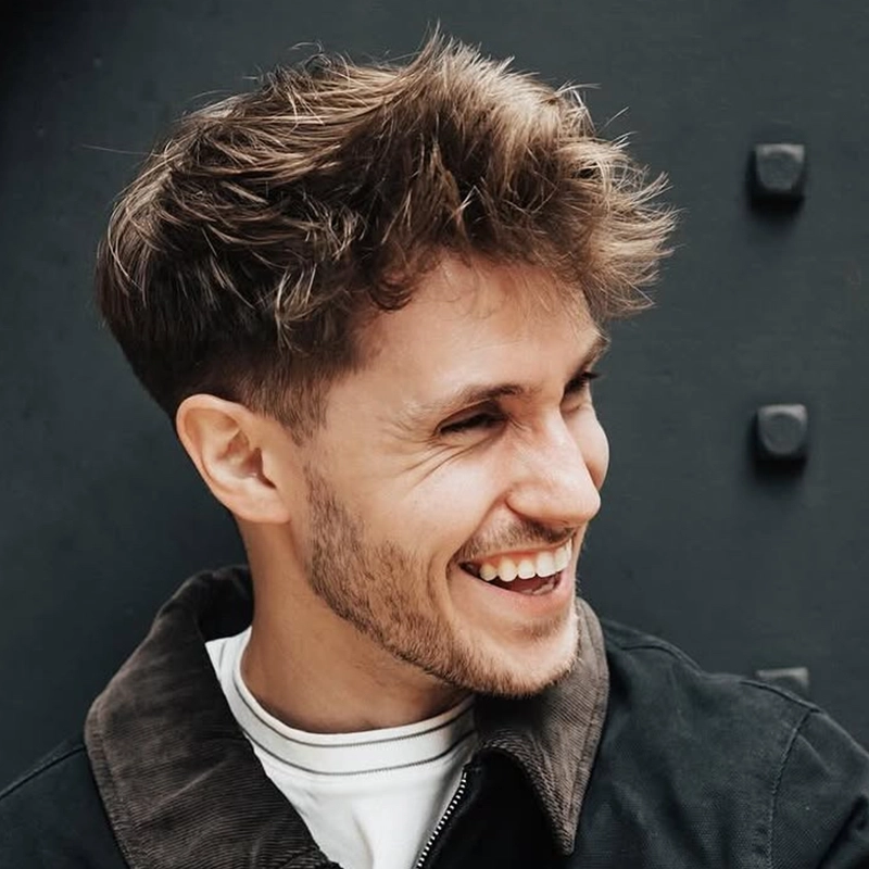 A young man with tousled brown hair and a short beard smiles, looking to the side. He wears a dark jacket over a white shirt. John Cardoza stands against a dark wall with large rivets in the background.