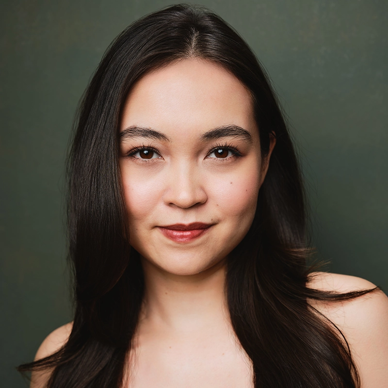 Katherine Winter, a young woman with long, dark hair and light skin, smiles slightly. She has brown eyes, natural makeup, and wears no jewelry. The muted dark green background and soft lighting create a warm, professional portrait.
