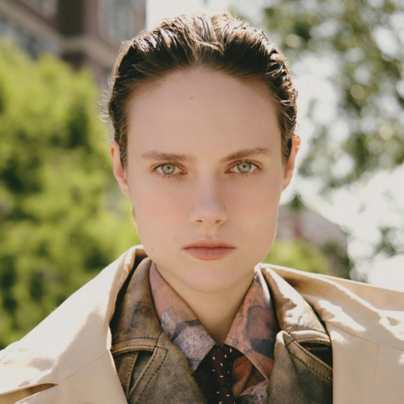 Katherine Winter, with light skin, slicked-back brown hair, and green eyes, stares seriously at the camera. She wears a tan coat over a patterned shirt and tie, set against blurred green foliage and buildings in natural light.