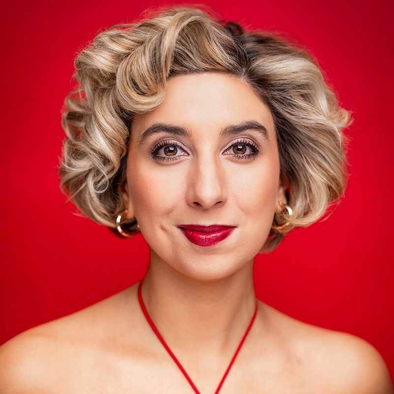 Katherine Winter, with short, wavy blonde hair and light skin, smiles gently against a bright red background. She wears bold red lipstick, hoop earrings, and a red halter strap, with subtle makeup and bare shoulders.