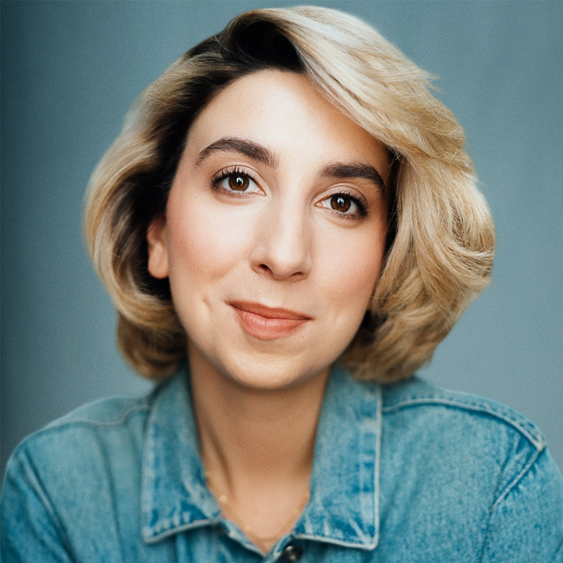 Katherine Winter, a woman with light skin and short, wavy blonde hair, smiles softly. She wears a light blue denim jacket and looks directly at the camera against a plain, muted blue background with soft, even lighting.