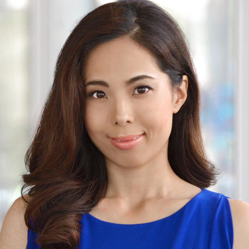 Kazue Kiyono, with long, wavy dark brown hair, smiles softly. She wears a sleeveless royal blue top against a light, blurred background. Her makeup is subtle and natural as she faces the camera directly.
