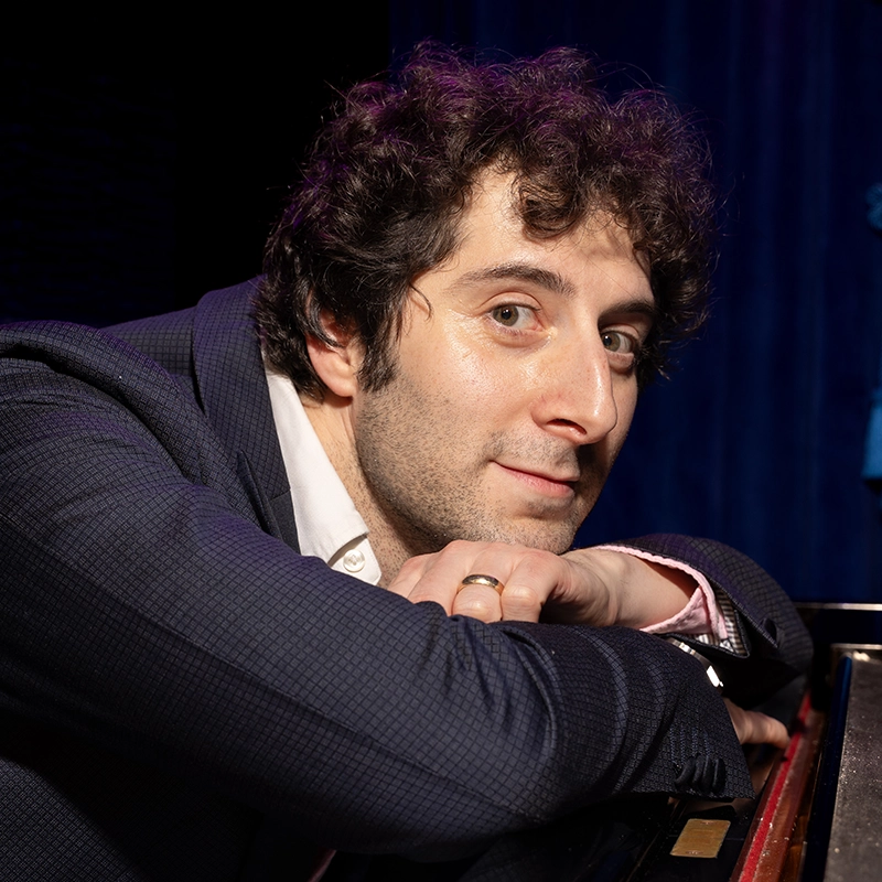 A man with curly dark hair leans on a piano, resting his arms and chin on it. He wears a dark blazer, light shirt, and a gold ring. He looks at the camera with a slight smile; behind him is Katherine Winter’s signature blue curtain and stage lighting.