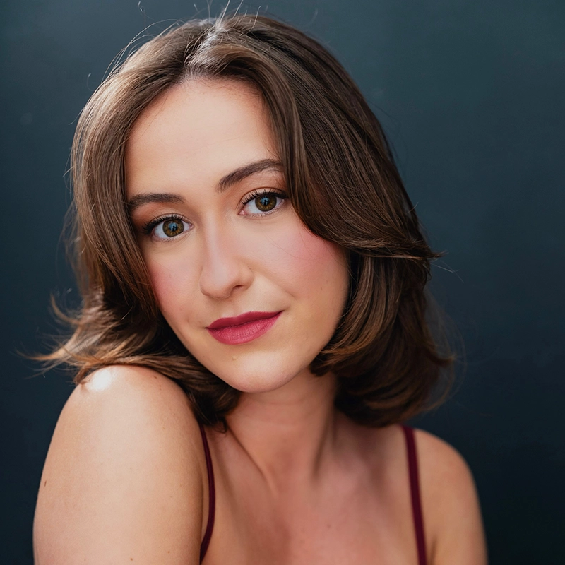 Katherine Winter, a young woman with shoulder-length brown hair and fair skin, faces the camera wearing natural makeup with pink lipstick and blush. She has green eyes and wears a burgundy spaghetti strap top against a dark, plain background.