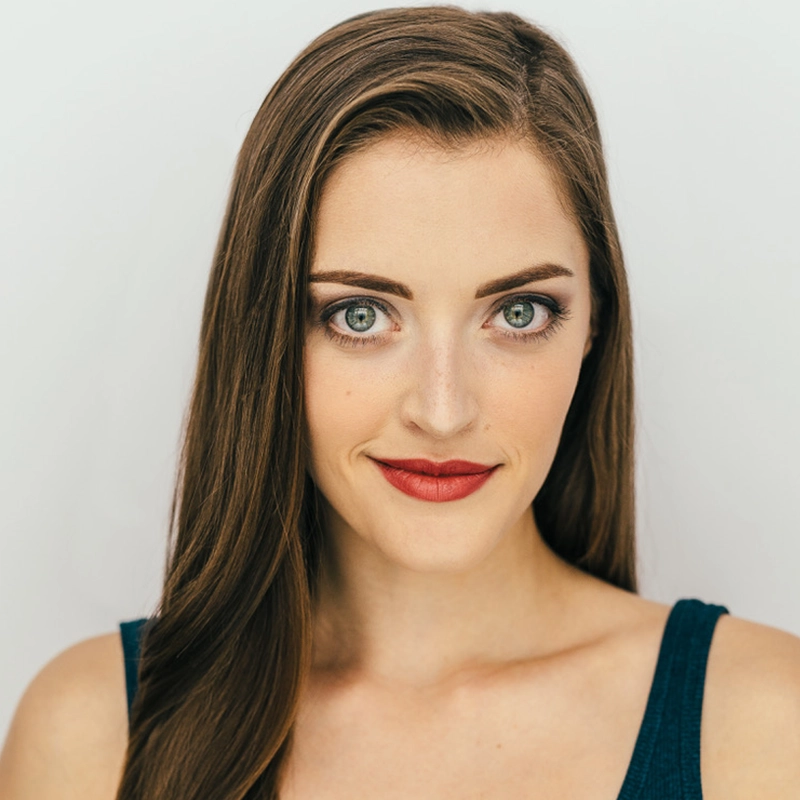 Katherine Winter, with long brown hair and green eyes, smiles softly. She wears a dark sleeveless top and red lipstick, with subtle makeup accentuating her features. The background is plain white as she faces the camera directly.