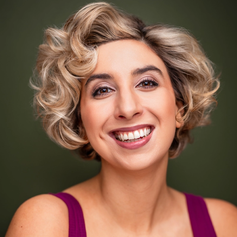 Katherine Winter, a young woman with short, curly blonde hair, smiles brightly at the camera. She wears a sleeveless magenta top and flawless, natural makeup against a solid olive green background, adding warmth to the portrait.