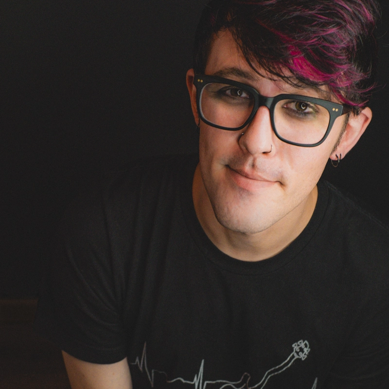 A young person with short dark hair and magenta highlights, wearing black glasses, a nose ring, and a black t-shirt, sits against a dark background, looking at the camera with a slight smile.