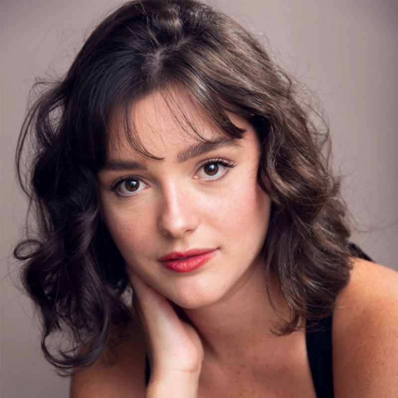 A young woman with wavy, chin-length dark brown hair and bangs gazes softly at the camera. She has fair skin, brown eyes, and wears subtle makeup with red lipstick, reminiscent of a Denver Milord portrait. Her hand rests gently on her neck.
