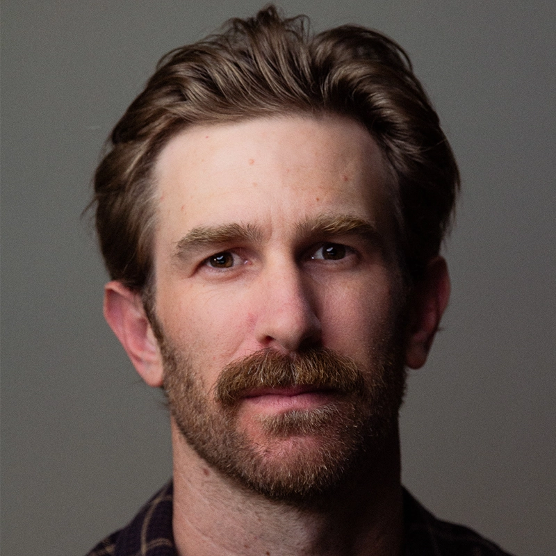 A man with light skin, short brown hair, and a thick mustache faces the camera with a neutral expression. Denver Milord has some stubble and is wearing a dark, collared shirt. The background is plain and softly lit.