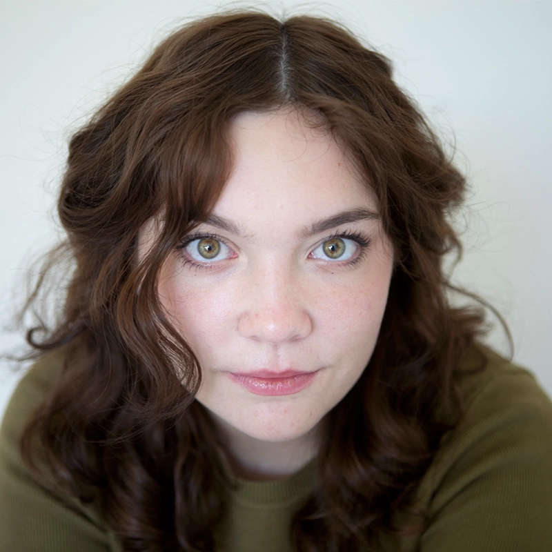 A young woman with wavy brown hair and green eyes looks directly at the camera. She has fair skin, minimal makeup, and is wearing an olive green top. The simple background keeps her face as the main focus in this Denver portrait.