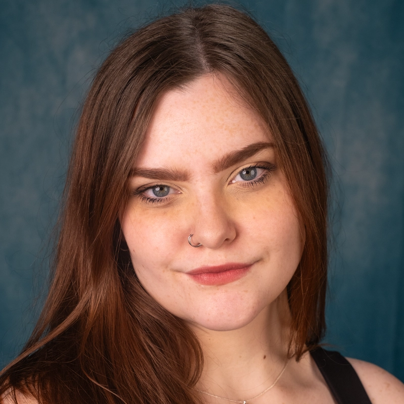 Young woman with long brown hair and fair skin looks at the camera with a slight smile. She has blue eyes, well-groomed brows, a small hoop nose ring, and wears a black top. The background is mottled dark blue.