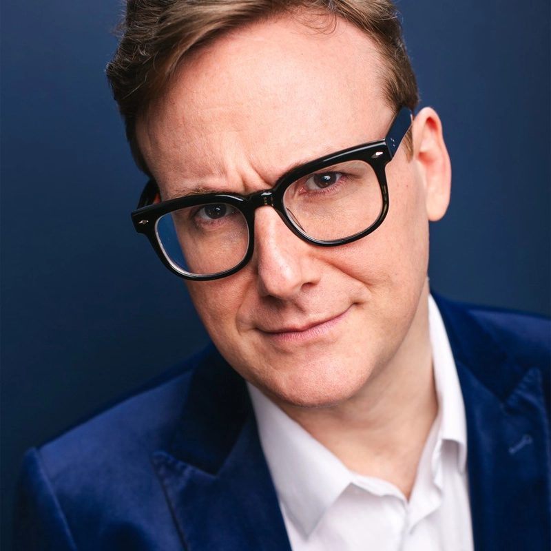 Abe Goldfarb, a man with short brown hair, wears black-rimmed glasses, a white shirt, and a dark blue blazer. He looks directly at the camera with a slight, inquisitive smile against a solid dark blue background.