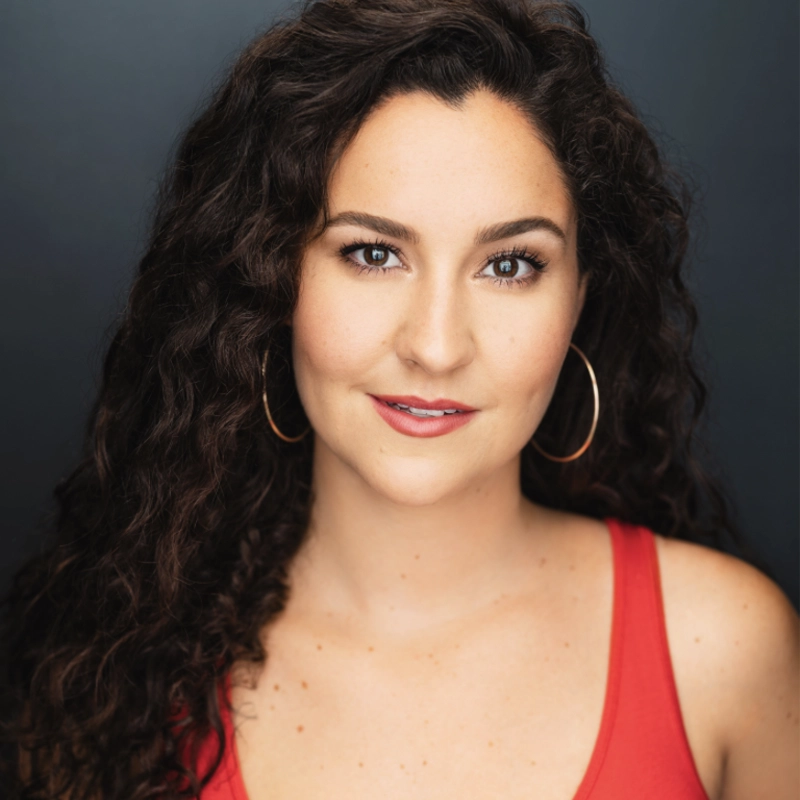 Sarah Hogewood, with long, dark curly hair and hoop earrings, looks at the camera in a sleeveless red top and subtle makeup. A slight smile graces her face as the dark, softly lit gradient background draws focus to her expression.