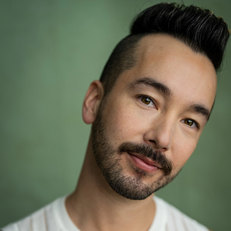 Keiji Ishiguri, with short dark hair styled upward and a trimmed beard, smiles gently. He wears a white shirt and looks slightly to the side against a soft green background, giving the portrait a friendly and approachable feel.
