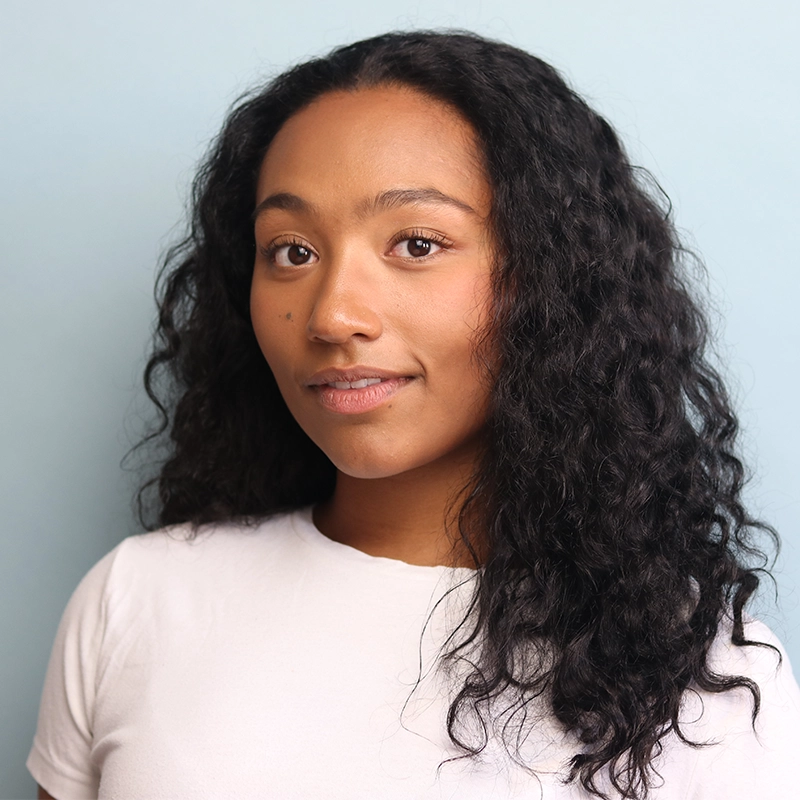 A young woman with medium brown skin and long, curly dark hair stands against a pale blue background in Denver. She is wearing a plain white t-shirt and has a calm, slight smile, looking directly at the camera.