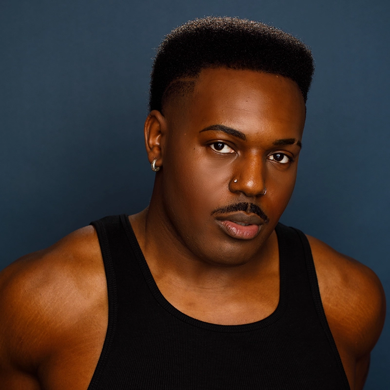 A person with dark skin, short afro high-top fade haircut, and a trimmed mustache looks directly at the camera. Denver Milord wears a black tank top, small earrings, and a nose ring, with a neutral expression against a dark blue background.
