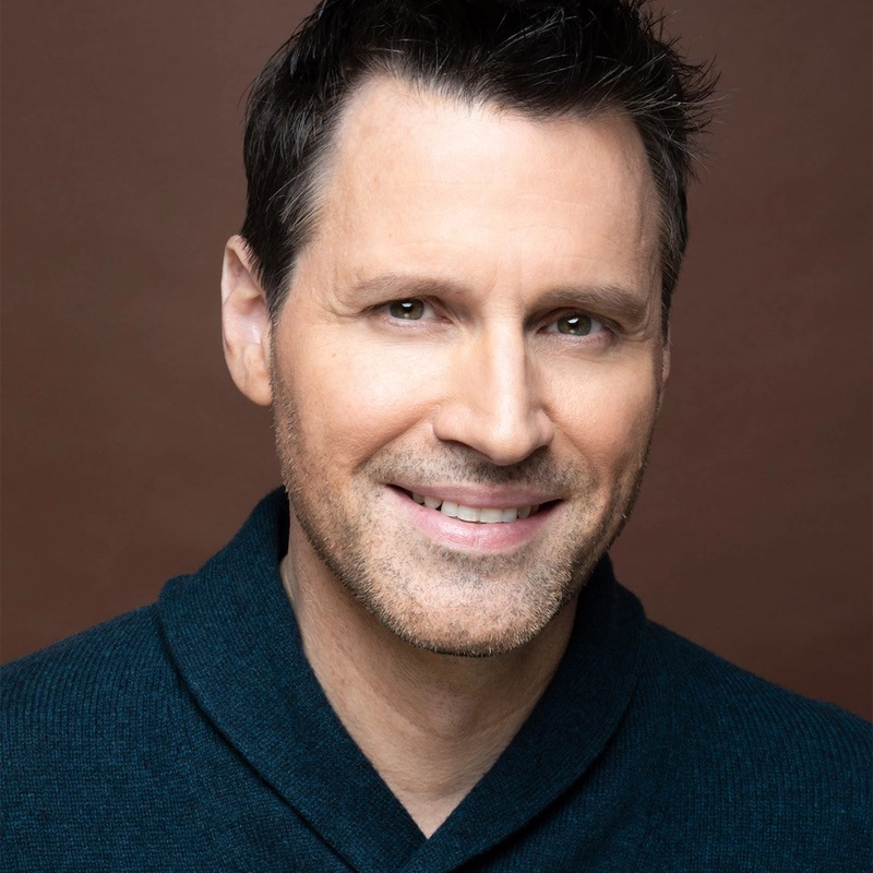 A middle-aged man, Patrick Oliver Jones, with short dark hair and light facial stubble smiles gently. He is wearing a dark blue sweater against a plain brown background, with soft lighting highlighting his approachable features.