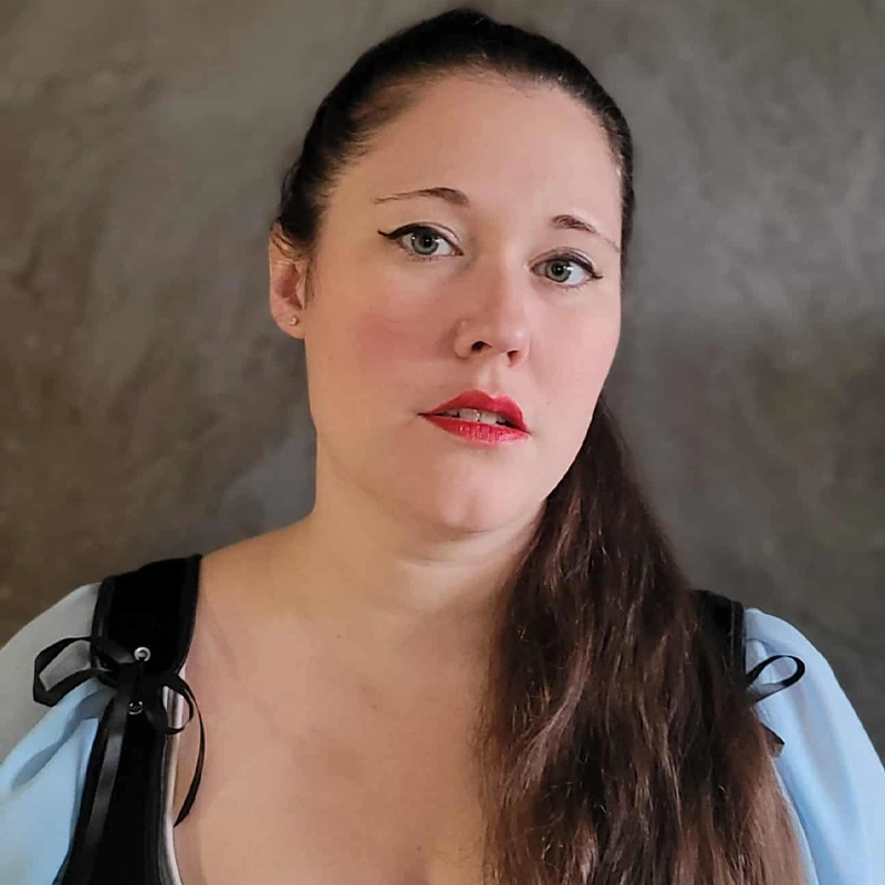 A woman with long brown hair pulled over one shoulder looks at the camera with a neutral expression. She wears winged eyeliner, red lipstick, a low-cut light blue top with black straps, and stands in front of a gray textured background.