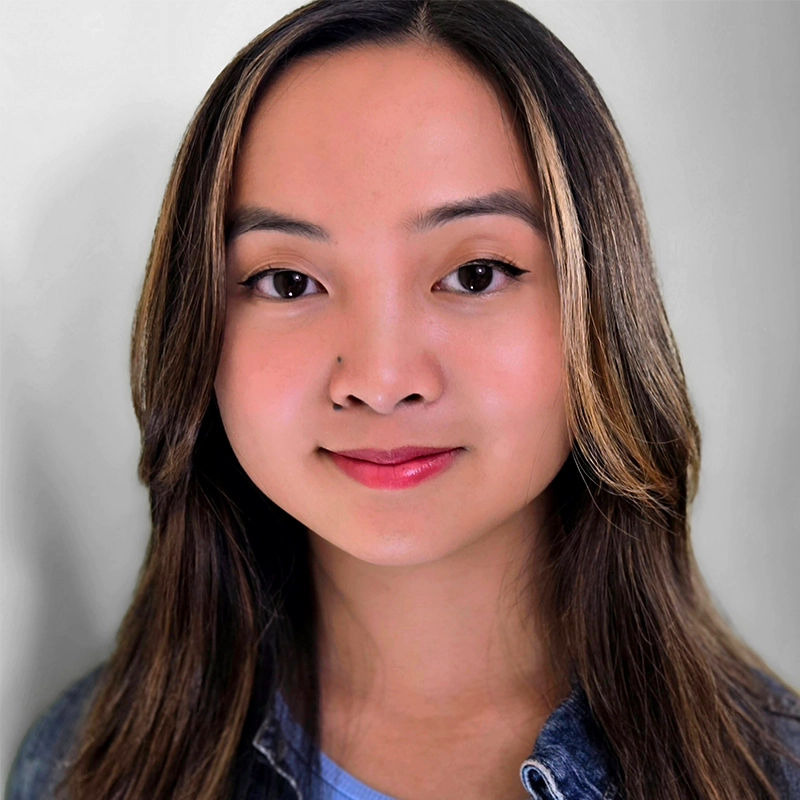 A young woman with long, straight brown hair and light highlights faces the camera, smiling gently. She has clear skin, brown eyes, and wears a blue top under a denim jacket. The background is softly blurred in light gray tones.