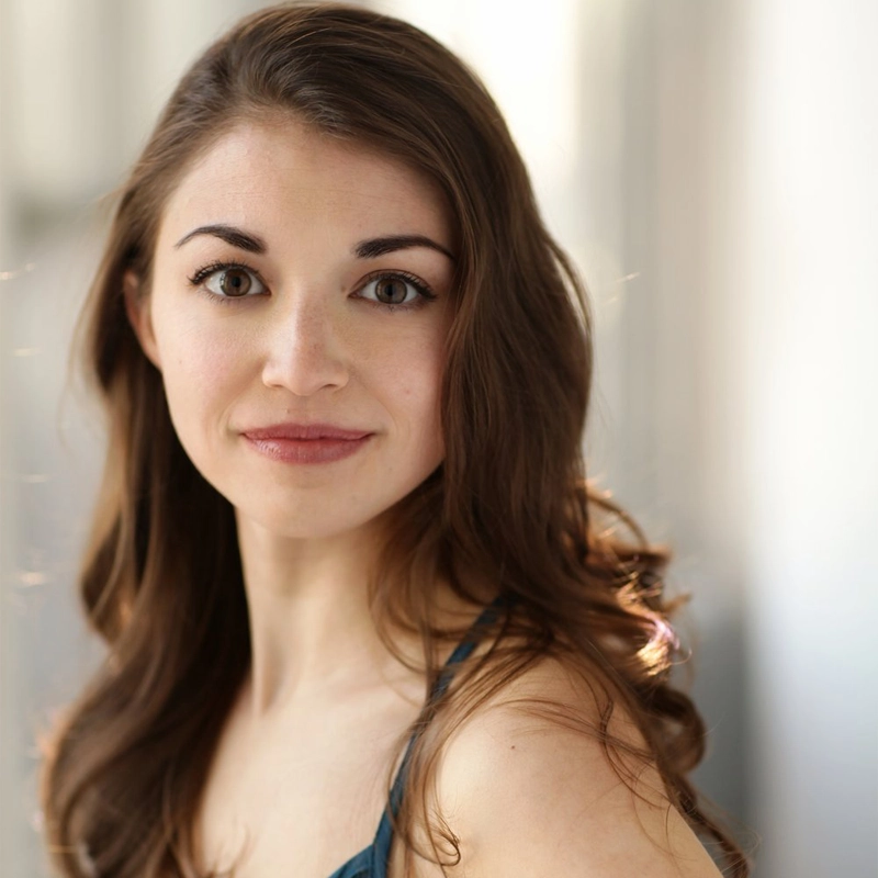 Beth DeMichele, a woman with long, wavy brown hair and fair skin, smiles softly at the camera. Wearing a sleeveless teal top against a blurred, light-filled indoor background, her gentle expression is enhanced by prominent eyebrows and clear eyes.