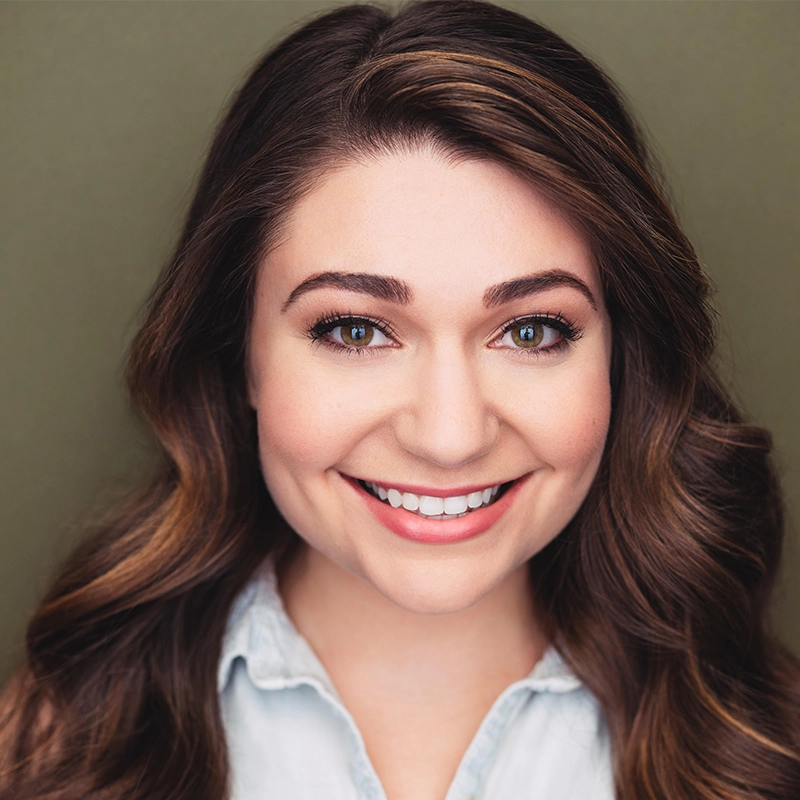 A young woman with long wavy brown hair smiles at the camera. She has light skin, brown eyes, and is wearing natural makeup. She is dressed in a light blue collared shirt. The background is a plain olive green.