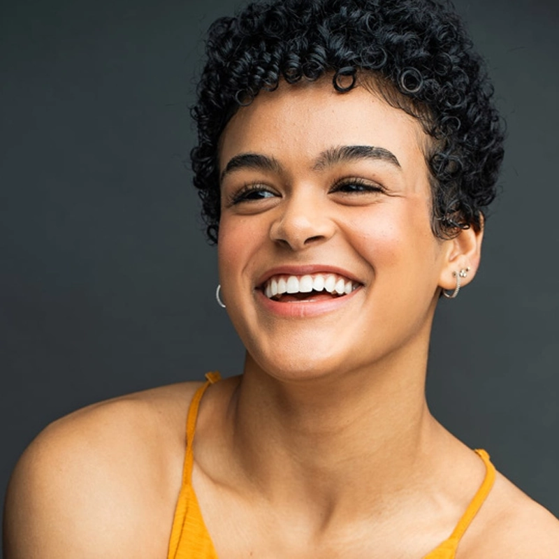 Gianna Harris, a young person with short, curly black hair, smiles brightly, showing their teeth. They wear a yellow sleeveless top and small hoop earrings. The dark gray background and lighting highlight their clear skin and joyful expression.