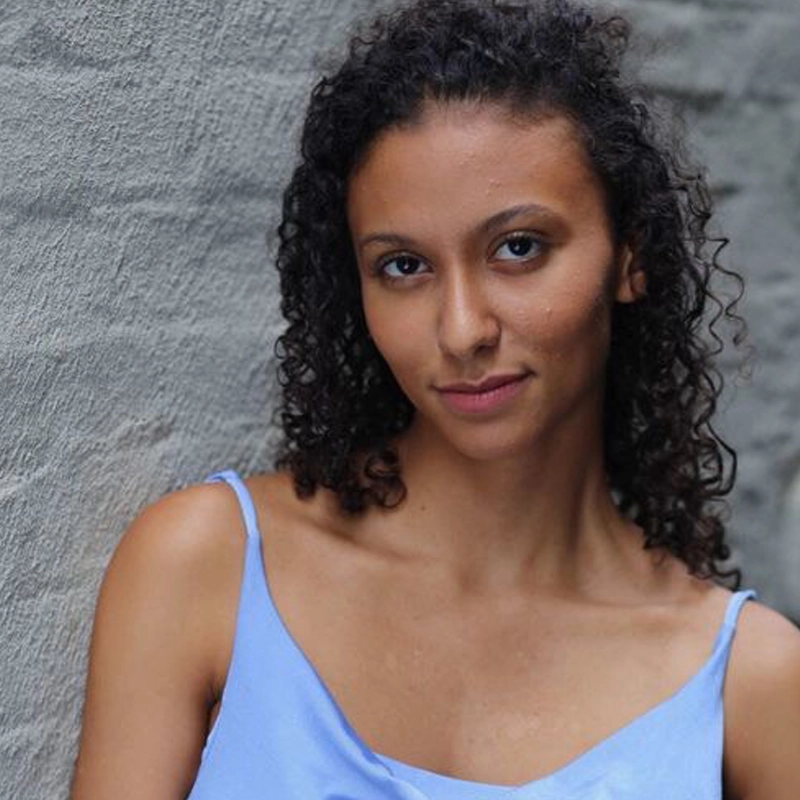 Joana Meurkens, with medium skin tone and curly dark hair, poses confidently in front of a textured gray wall. She wears a light blue spaghetti strap top and gazes at the camera with a gentle, slight smile.