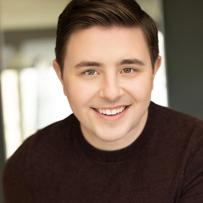 Jake Nicholson, a young man with short brown hair and hazel eyes, smiles at the camera. He is wearing a dark brown sweater and photographed in soft natural light, with a blurred indoor background that adds to the warm, approachable mood.