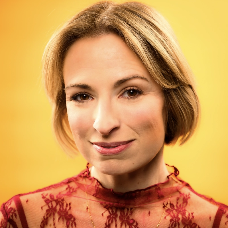 Stephanie Lazard, with short blonde hair, smiles gently. She wears a sheer red lace top and a thin necklace. The bright, solid yellow background gives the image a warm, vibrant feel as lighting highlights her face and hair.