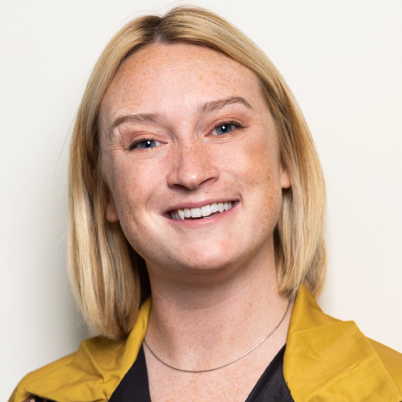 A smiling person with fair skin, straight blonde hair in a chin-length bob, and freckles. They are wearing a yellow jacket over a black top and a thin silver necklace, posed against a light background, reminiscent of Kaila Mullady’s vibrant style.