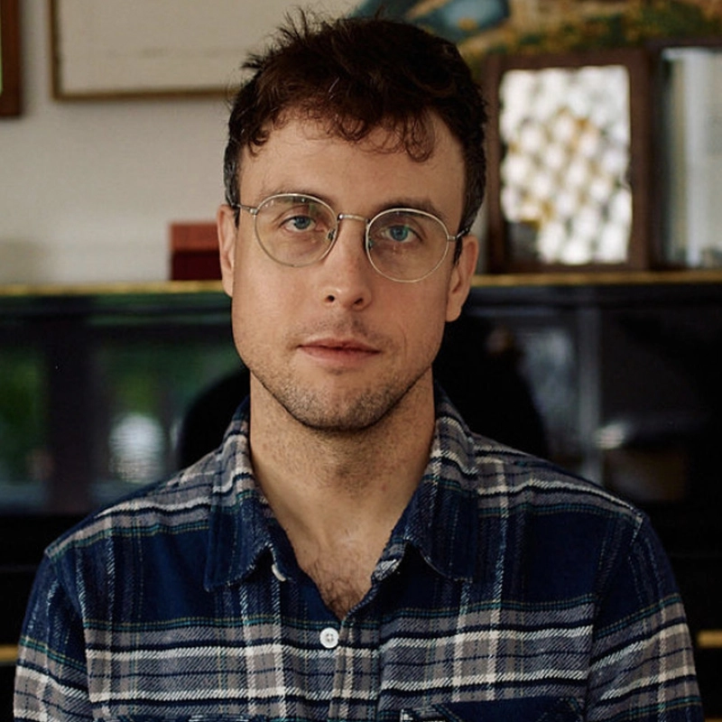 Mark Sonnenblick is a young man with short brown hair, light stubble, and thin-rimmed round glasses, wearing a dark blue and white plaid button-down shirt. He looks directly at the camera with a neutral expression in an indoor setting with a blurred background.