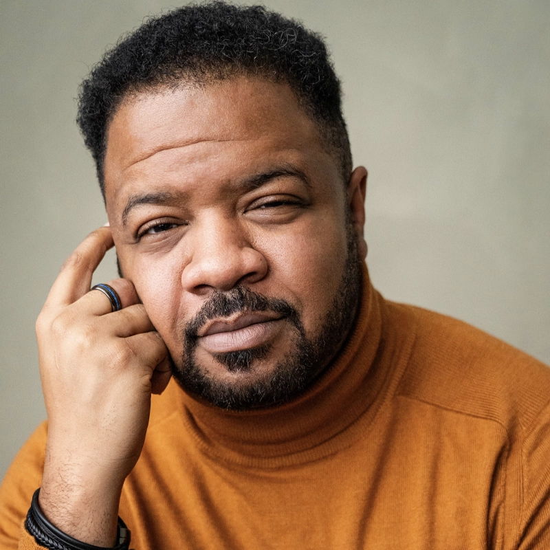 A man with short curly hair and a neatly groomed beard wears a burnt orange turtleneck sweater. Michael Andreaus rests his face on his left hand, looking thoughtfully at the camera against a plain, soft green background.