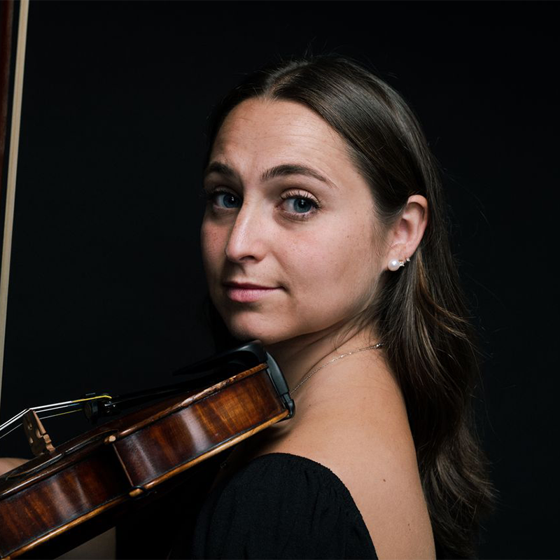 Hannah Cohen, with long brown hair and blue eyes, gazes at the camera, holding a violin under her chin. She wears a black top and pearl earrings, softly lit against a dark background that highlights her face and the violin.