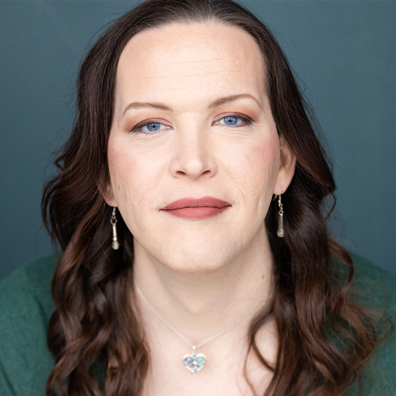 Khaila Wilcoxon, with long brown wavy hair, blue eyes, and fair skin, looks directly at the camera. She wears subtle makeup, drop earrings, and a heart-shaped pendant necklace against a solid muted blue-gray background.