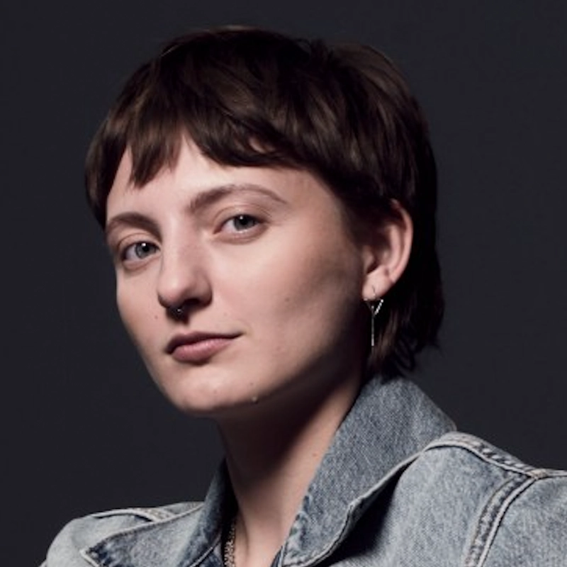 Ariana Notartomaso, with short brown hair and fair skin, gazes confidently at the camera. She sports a septum ring, a silver earring, and a light denim jacket over a dark top, set against a plain dark background that highlights her expression.