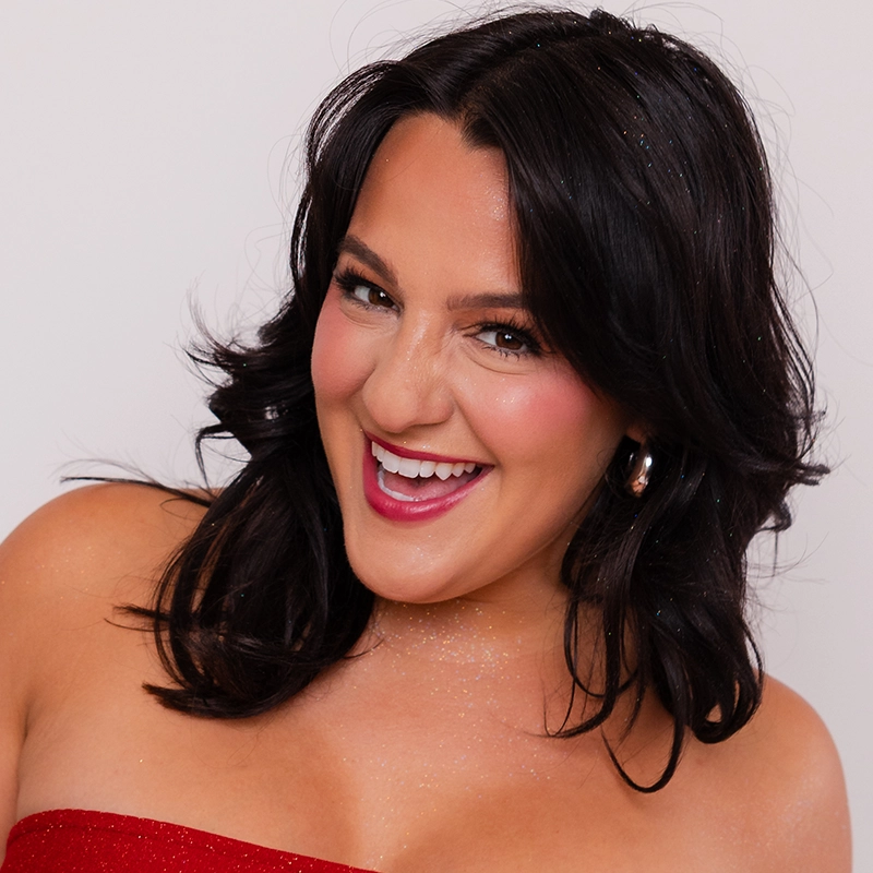 Khaila Wilcoxon, with medium-length dark hair, smiles widely in a strapless red top and gold hoop earrings. Glitter accents her skin and hair, while pink blush and lipstick complete her look against a plain white background.