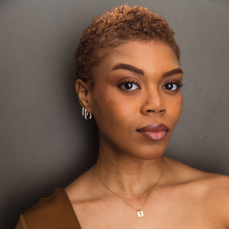 A woman with short curly hair, resembling Khaila Wilcoxon, looks at the camera in a brown off-the-shoulder top, small hoop earrings, and a gold square pendant. The background is plain gray, and she has a calm, confident expression.