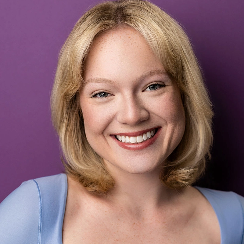 A smiling woman with short blonde hair and fair skin wears a light blue top. Khaila Wilcoxon faces the camera in front of a solid purple background. The lighting is bright, highlighting her clear complexion and cheerful expression.
