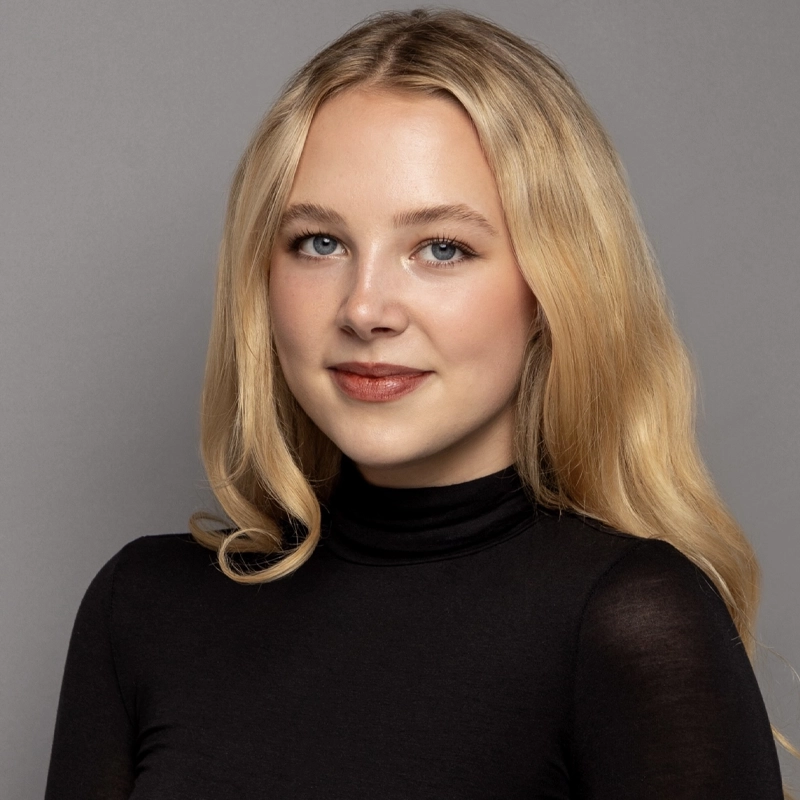 Lauren Robinson, a young woman with long blonde hair and blue eyes, wears a black turtleneck. She is smiling softly and poses in front of a plain gray background. Her expression is warm and confident, and her makeup is natural.