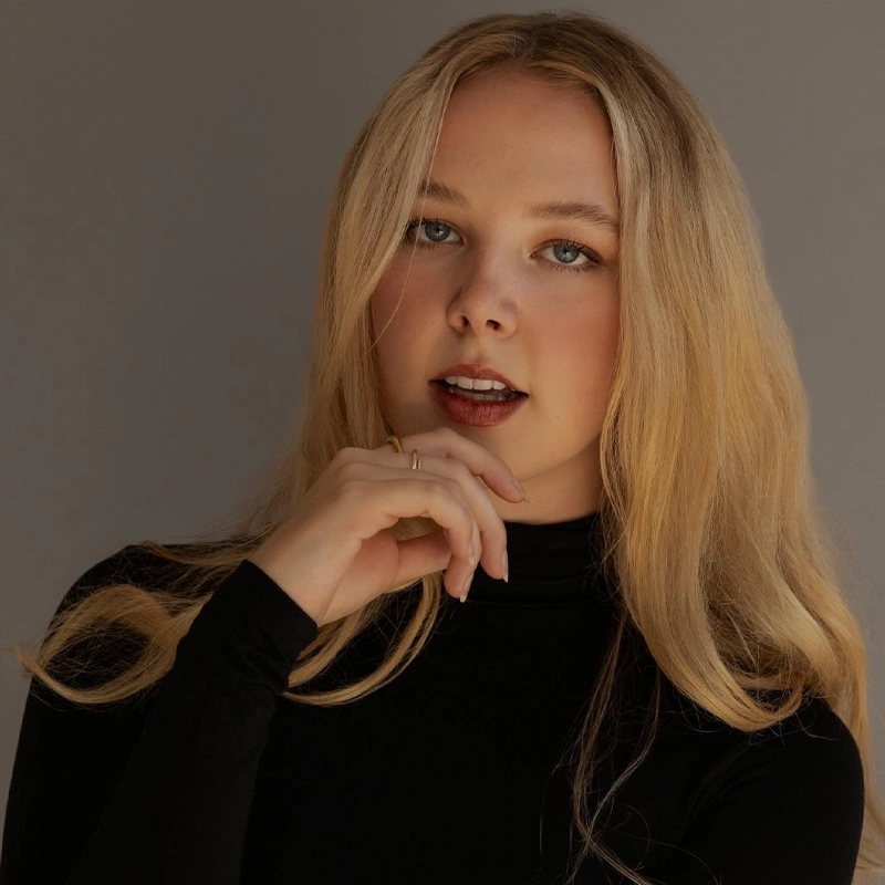 Lauren Robinson, a young woman with long blonde hair and blue eyes, poses against a neutral background. She wears a black turtleneck and gazes at the camera with her mouth slightly open, lightly touching her chin with her left hand in soft, warm light.