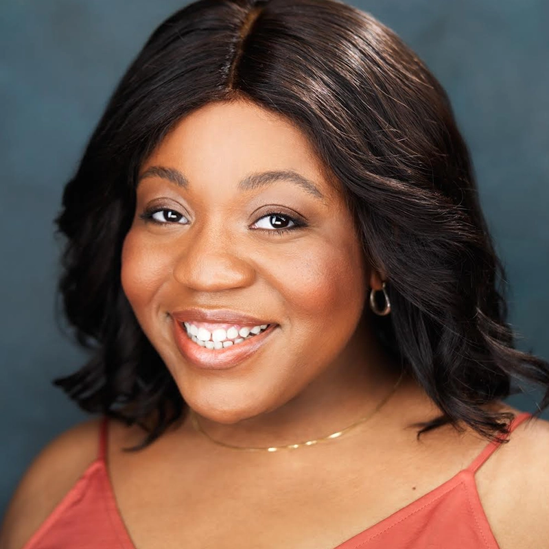 Khaila Wilcoxon, with shoulder-length wavy dark hair, smiles warmly at the camera. She wears a rust-colored top, a delicate gold necklace, and small hoop earrings. The soft blue background highlights her cheerful expression.