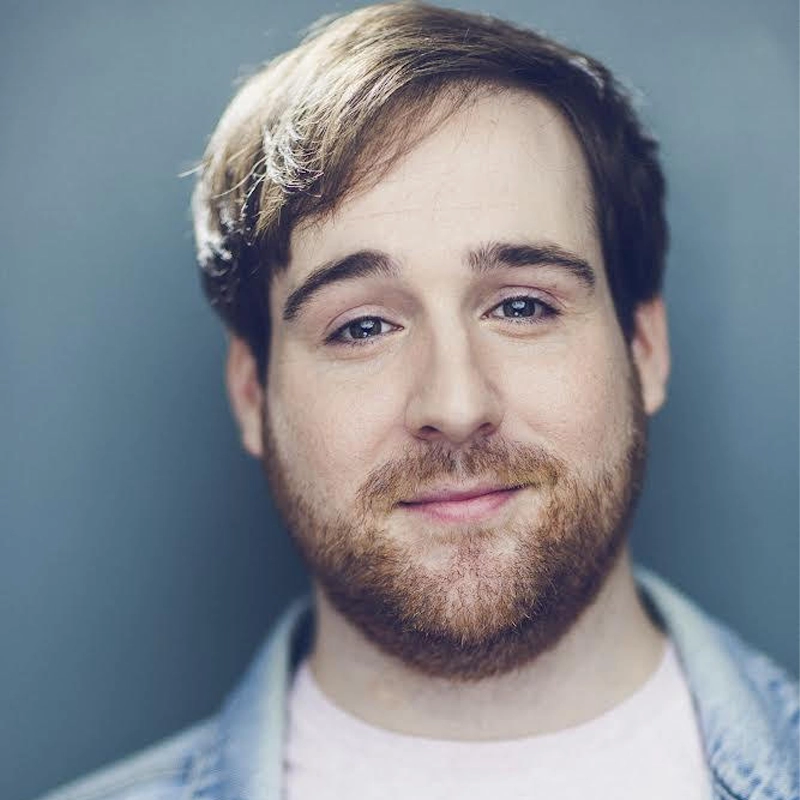 A young man with short brown hair and a full beard looks at the camera, smiling slightly. He wears a light denim jacket over a pale shirt. The background is softly blurred in shades of blue and gray, and the lighting is gentle and even—capturing Liam Fennecken in a relaxed, approachable moment.